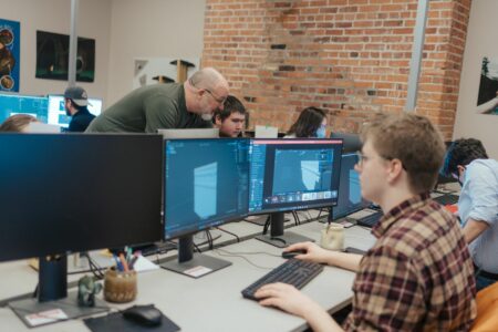 A NBCCD student sits at a desk in the 3D Digital Design classroom. He works with two monitors, making 3d objects on the screen. A instructor is helping out another student at a computer in the background.