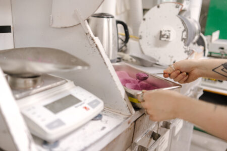 A person measuring vibrant pink powder with a spoon in a ceramics studio, beside a digital scale and equipment.