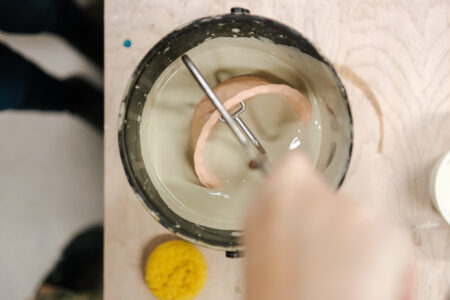A ceramic piece being dipped into a bucket of glaze using metal tongs, with a sponge nearby on the work table.
