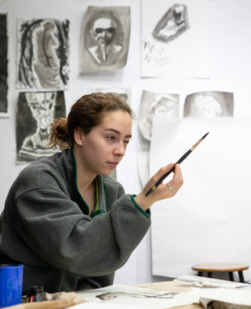 A student looks at the subject she is painting. Other sketches in charcoal are on the wall behind her.