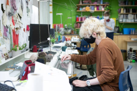 A student uses a knitting machine in the Textiles Studio on NBCCD's campus. They are following campus COVID-19 guidelines by wearing a mask.