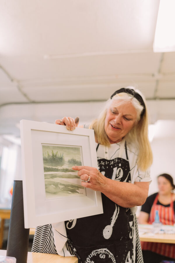 An instructor in the Honeybee Folk School teaches how to abstract water colour paint. She points to a water colour painting with students in the background regarding her intently.