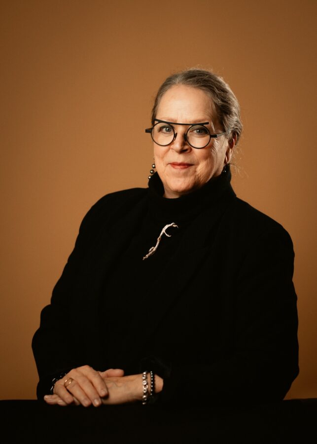 An artistic woman wearing all black with glasses sitting for the camera with a burnt orange backdrop.