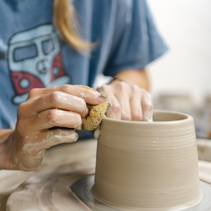 Close-up of hands covered in wet clay using a sponge to smooth the surface of a clay vessel on a spinning pottery wheel.